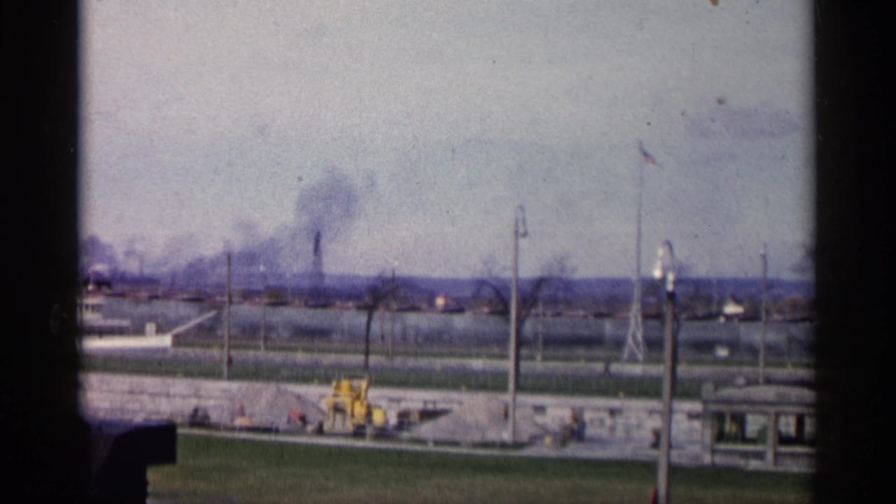 Industrial smoke plume seen beyond a road, wall, flagpole, and yellow tractor.