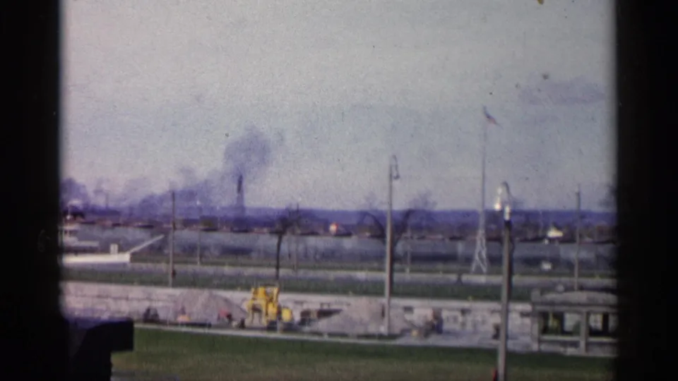 A faded 8mm view of Michigan industrial smoke in 1960, framed across a road or waterfront with a yellow tractor, flagpole, utility poles, and low buildings.