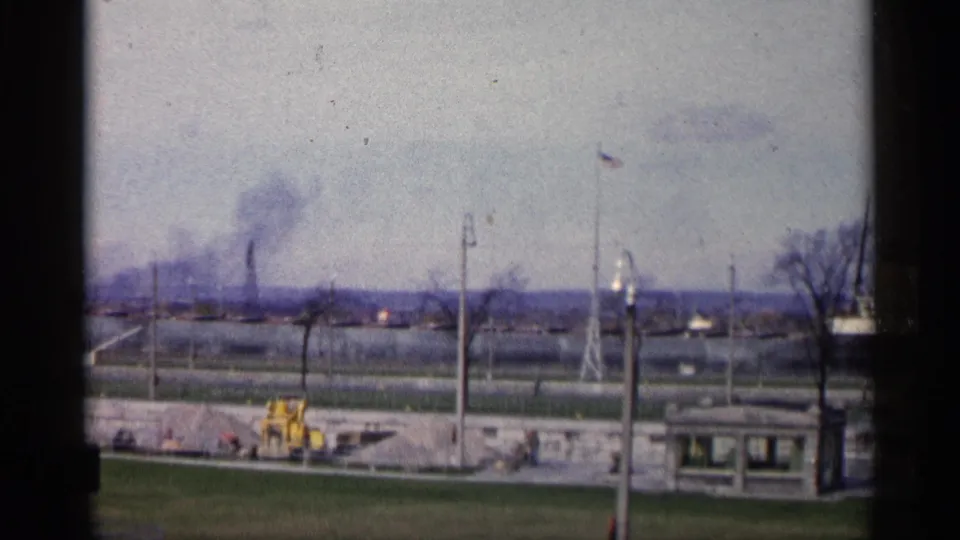 1960 Michigan home-movie frame with distant factory smoke, utility poles, a yellow tractor near a wall, grass, and a small roadside structure at lower right.