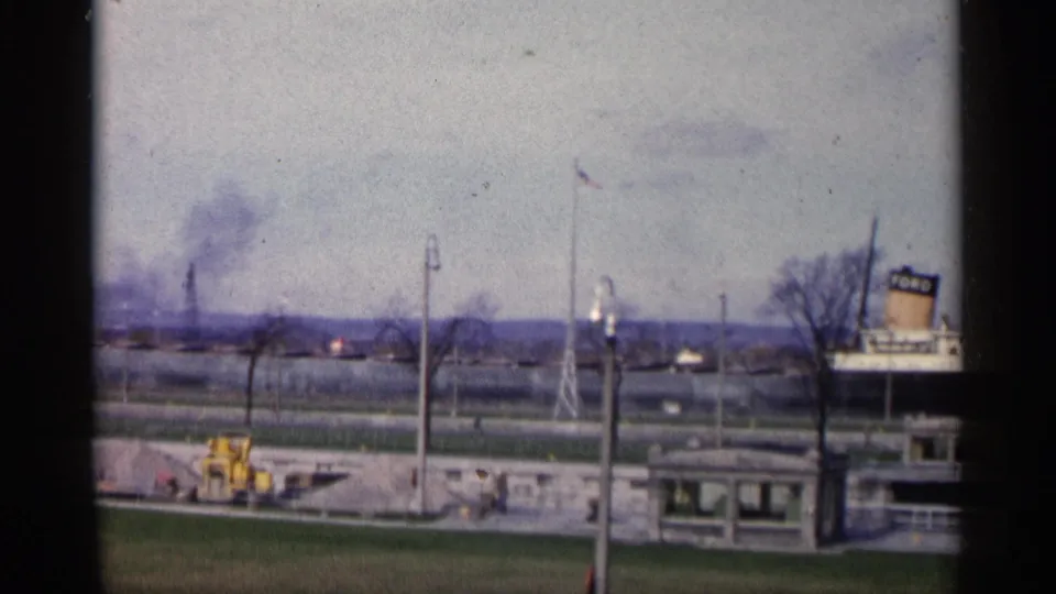 Final 1960 Michigan 8mm frame showing smoke over an industrial area, a yellow tractor, flagpole, utility poles, small roadside building, bare tree, and partial sign.