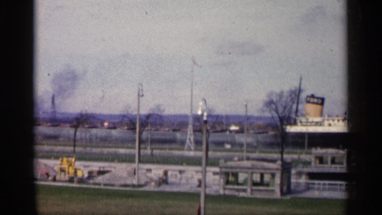 1960 Michigan home-movie frame showing a docked ship with a FORD-marked smokestack near a pier, flagpole, bare trees, and grassy foreground.