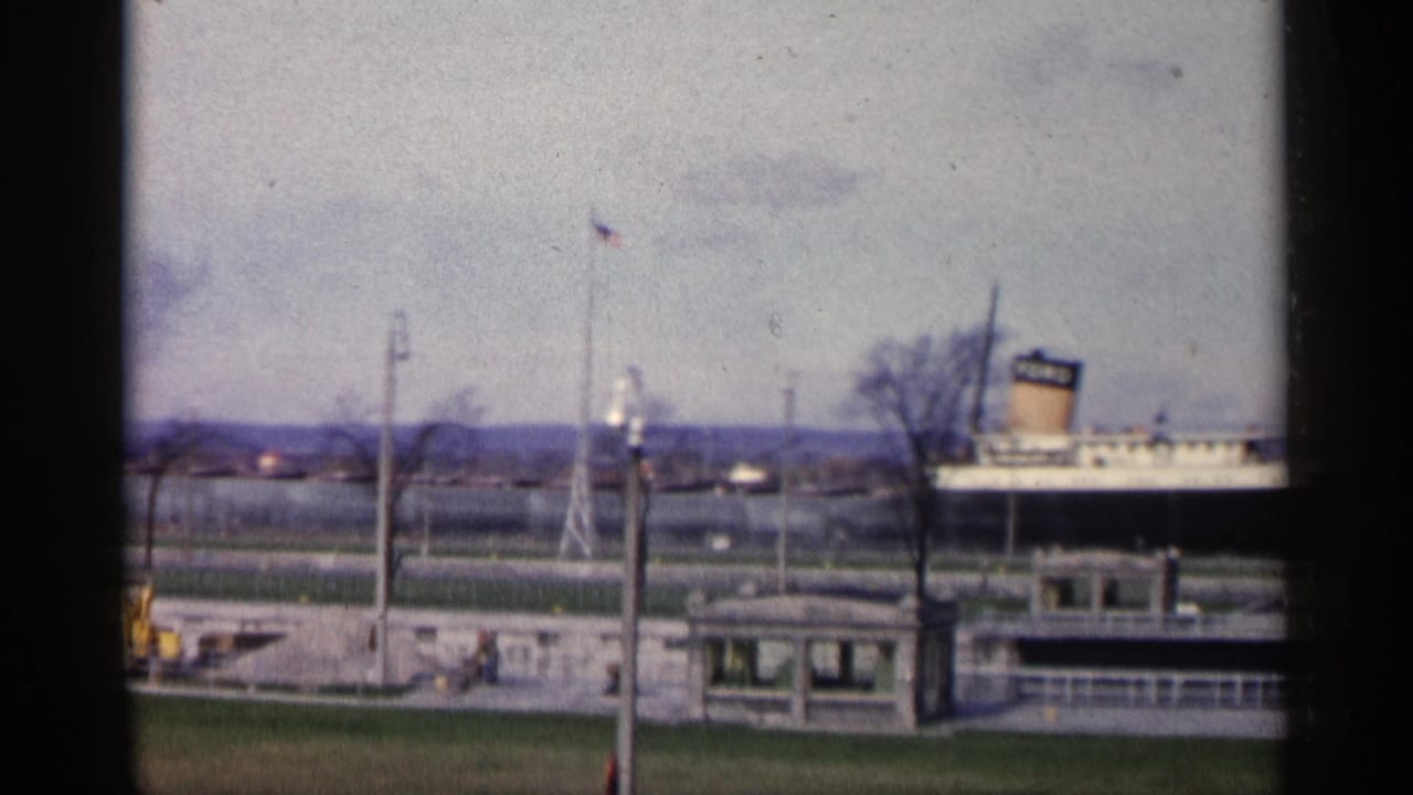 1960 Michigan waterfront frame with the FORD-marked ship partly visible behind a pier, American flag, lamp posts, bare trees, and small structure.