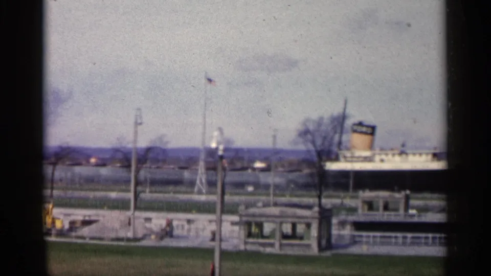 1960 Michigan waterfront frame with the FORD-marked ship partly visible behind a pier, American flag, lamp posts, bare trees, and small structure.