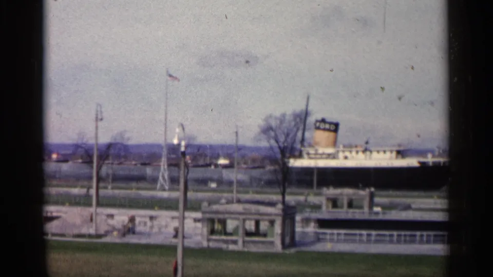 1960 Michigan home-movie frame showing the docked FORD-marked ship, flagpole, stone pier, bare tree, waterfront building, and road edge.