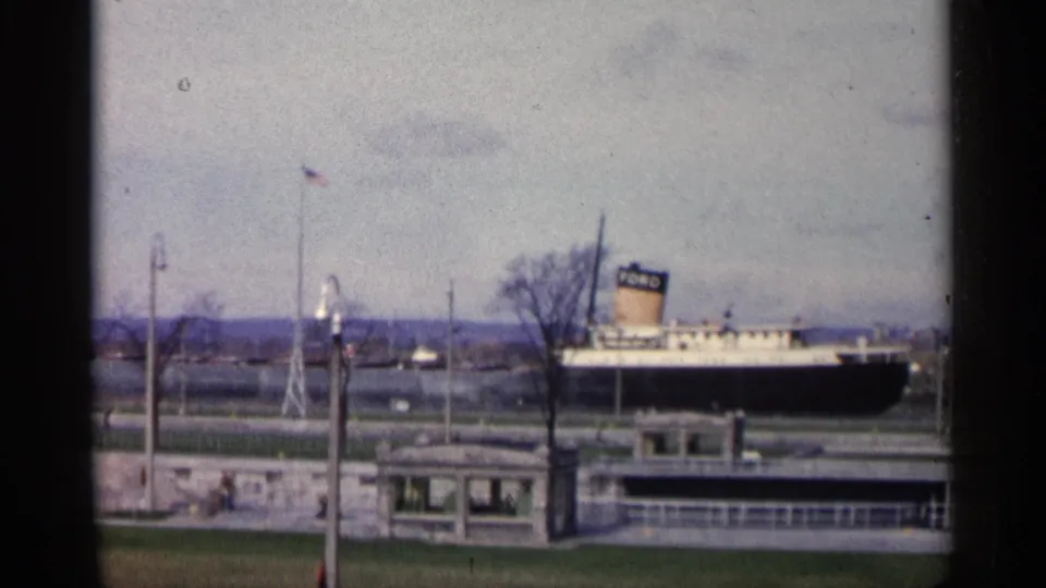 1960 Michigan frame with the ship more centered behind the waterfront building, stone pier, flagpole, bare trees, and muted shoreline beyond.