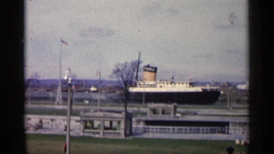 1960 Michigan 8mm frame showing a black Ford-marked ship docked beside a canal lock, with grassy banks and waterfront structures.