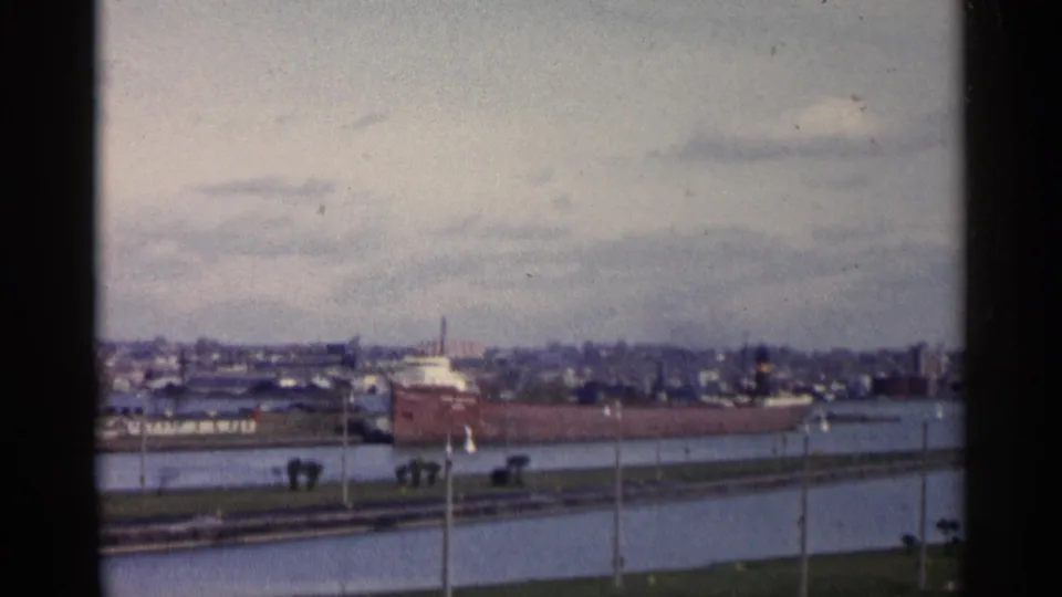 1960 Michigan 8mm frame showing a red freighter along the canal, with water, shoreline poles and distant industrial buildings.