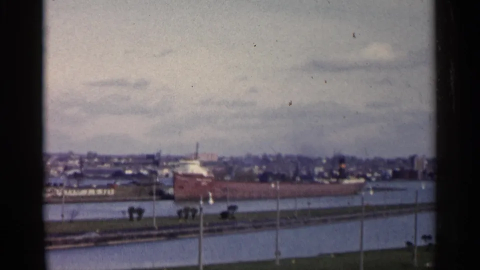 1960 Michigan 8mm frame of a red freighter across the canal with shoreline infrastructure and a distant industrial skyline.