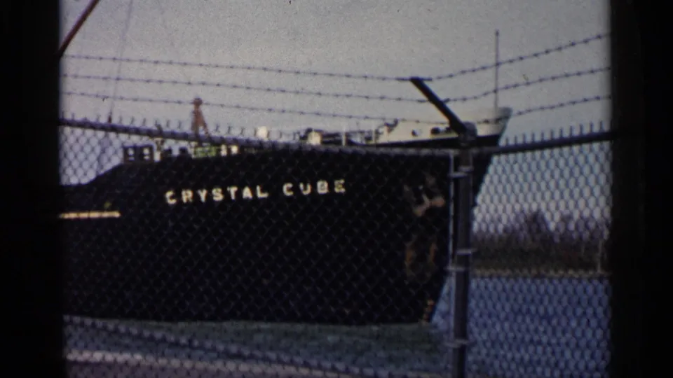 1960 Michigan 8mm frame showing the dark-hulled tanker Crystal Cube docked behind a chain-link fence with barbed wire.