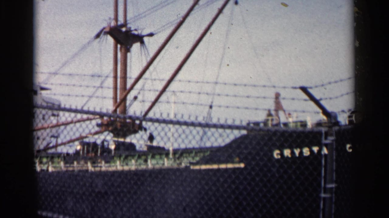 1960 Michigan 8mm frame of a large vessel viewed through a chain-link fence, showing mast rigging, hull lettering, and dockside barriers.