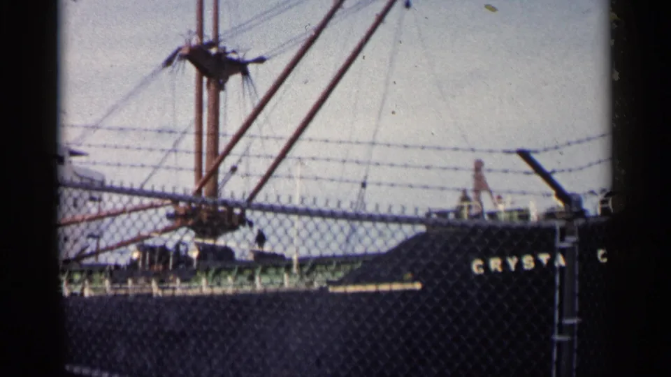 1960 Michigan 8mm frame of a large vessel viewed through a chain-link fence, showing mast rigging, hull lettering, and dockside barriers.