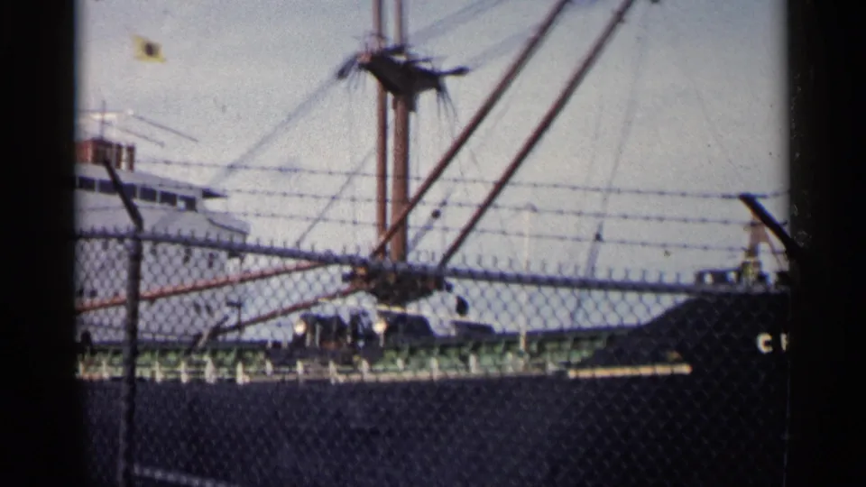 1960 Michigan 8mm frame showing the vessel behind a security fence, with its white superstructure, dark hull, and rigging partially obscured.