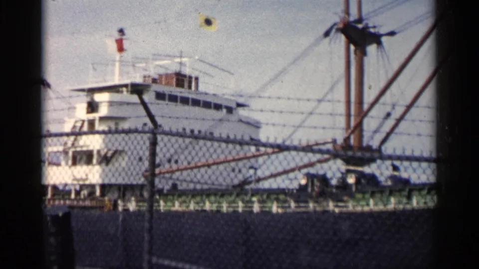 1960 Michigan 8mm frame of the vessel's white cabin and mast rigging viewed from behind a dockside chain-link fence.