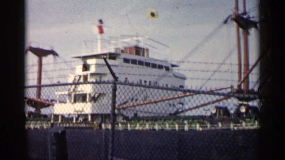 1960 Michigan 8mm frame showing a large white docked vessel behind a chain-link fence under a bright sky.
