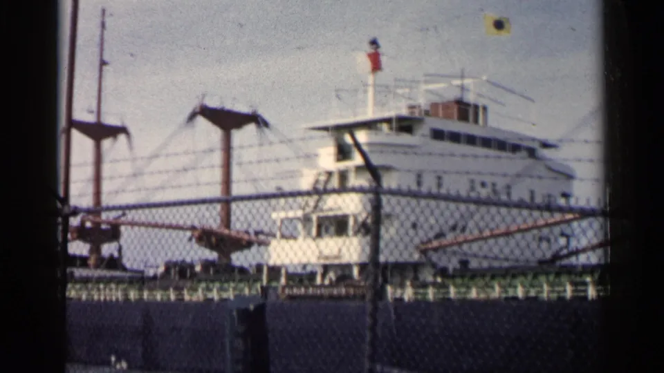 A large white vessel remains docked behind fencing, with upper-deck windows, railings, and port rigging visible.