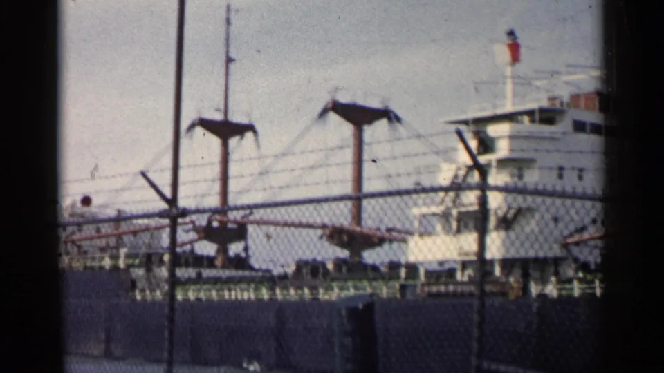 The handheld view shifts along the docked vessel, showing masts, rigging, fence mesh, and part of the white superstructure.