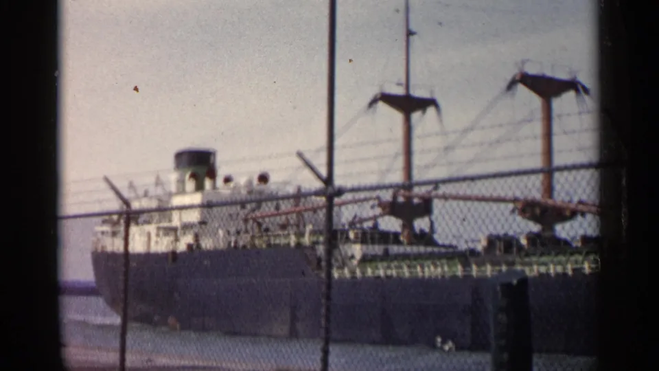 1960 Michigan 8mm frame showing a dark-hulled cargo ship with white upper decks viewed through a chain-link fence.
