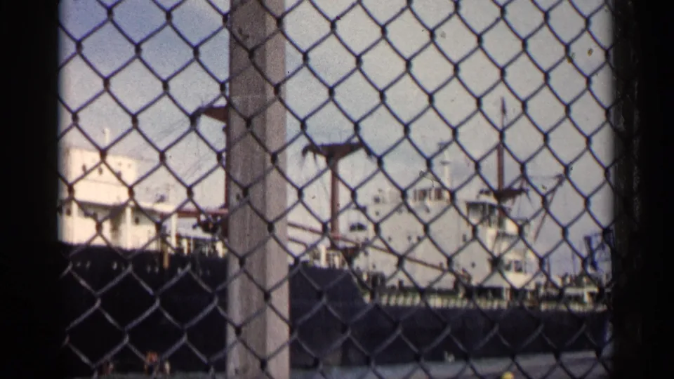 A closer 8mm view shows the freighter's white deck structures and cranes behind a chain-link fence and central post.