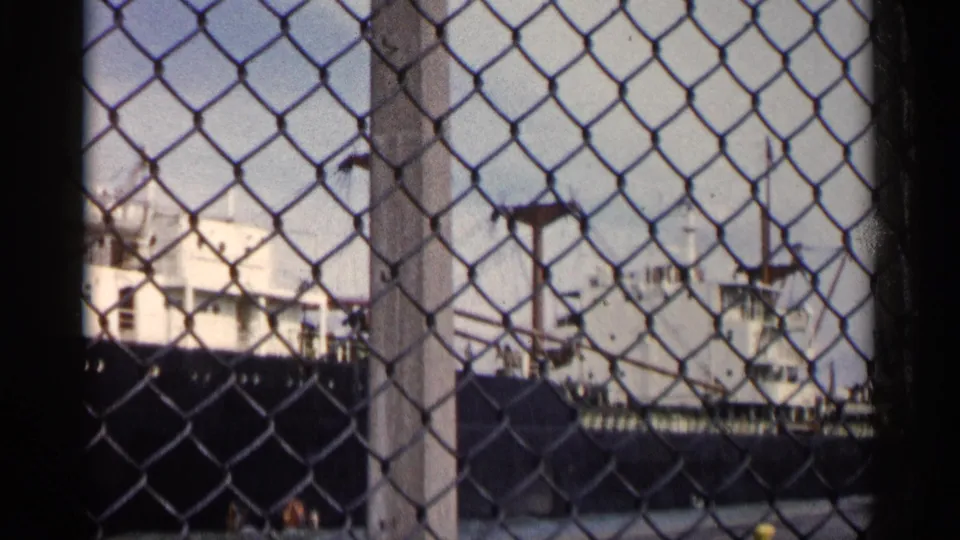 The final frame holds on the docked Michigan cargo ship, its white superstructure and cranes filtered through the foreground fence.