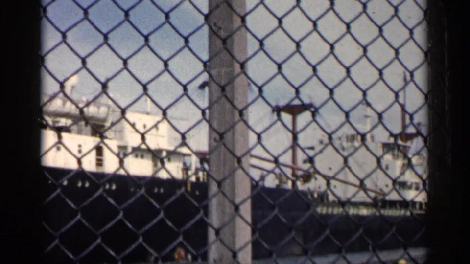 1960 Michigan home-movie frame of a large white ship with a dark hull viewed through chain-link fencing.