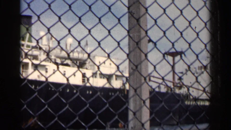 A docked ship appears beyond chain-link fencing, with white decks and a dark hull under a bright sky.