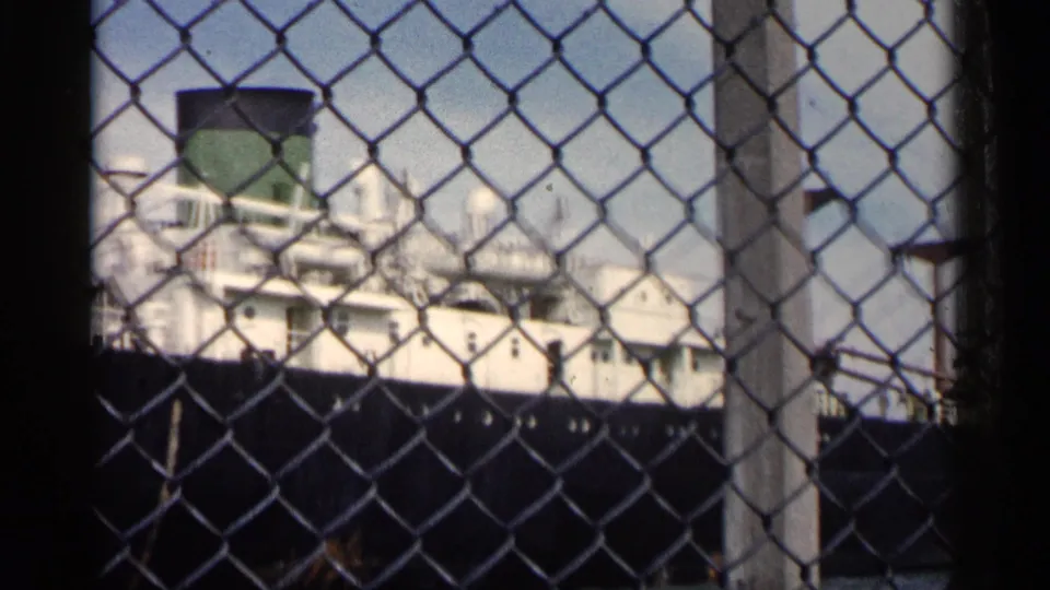 The ship's white upper decks and green smokestack are visible behind a chain-link fence in the Michigan port footage.