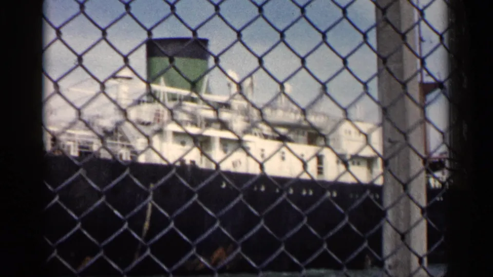 Final frame shows the ship's green smokestack, white superstructure, and dark hull behind waterfront fencing.