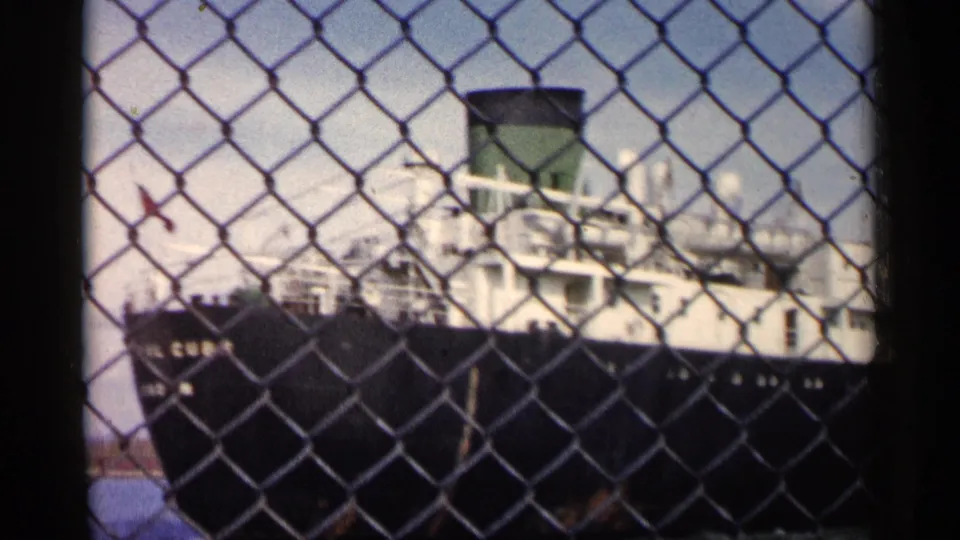 1960 Michigan 8mm frame of a large ship partially obscured by chain-link fencing along a waterfront.