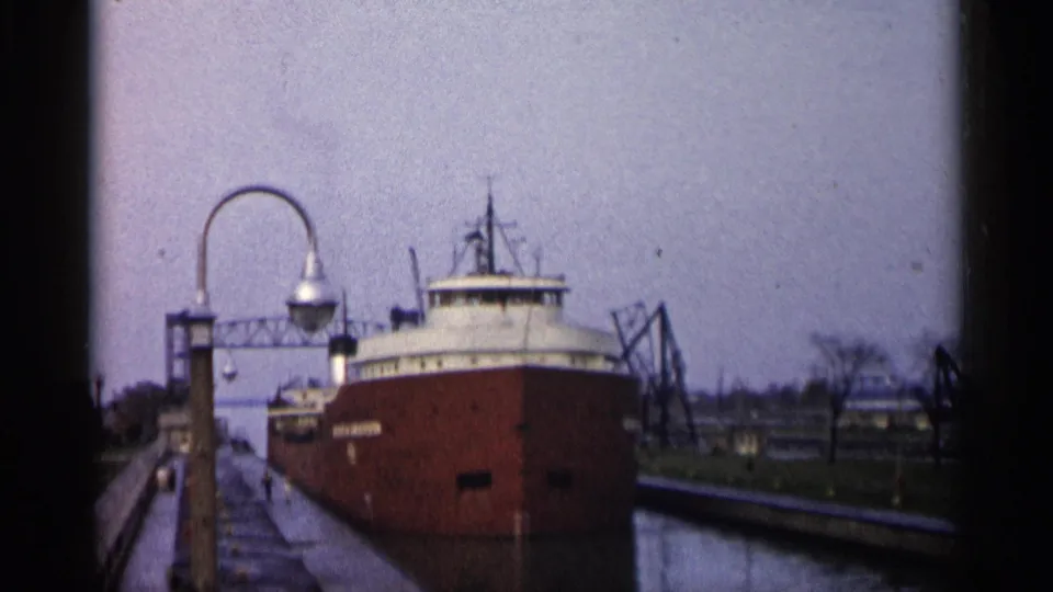 Handheld 8mm view of a red freighter in a Michigan lock channel with dock lighting and harbor equipment nearby.