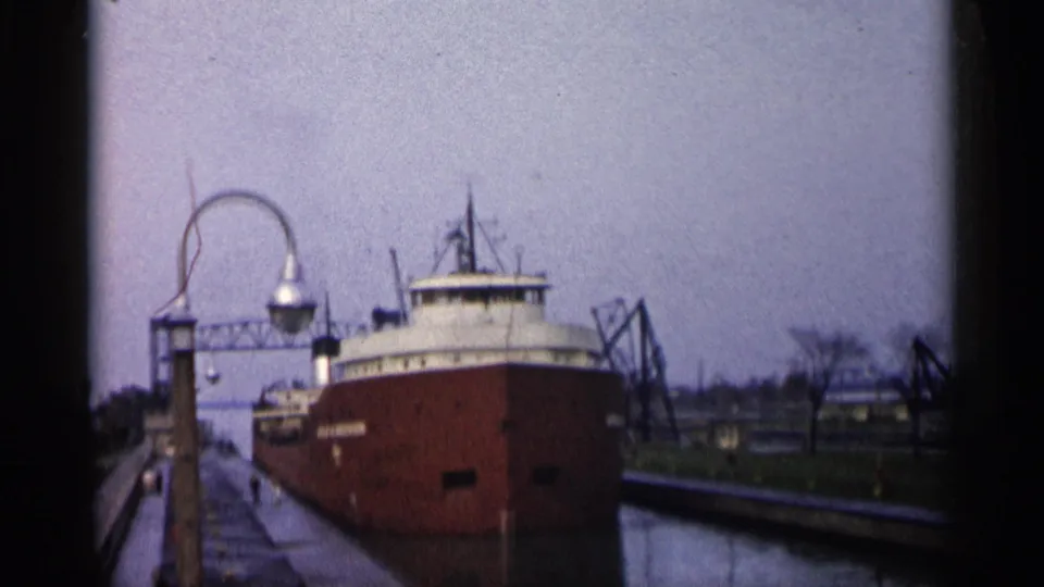 1960 Michigan harbor frame showing the red freighter in a lock, bordered by water, dock walls, a lamppost, and industrial structures.