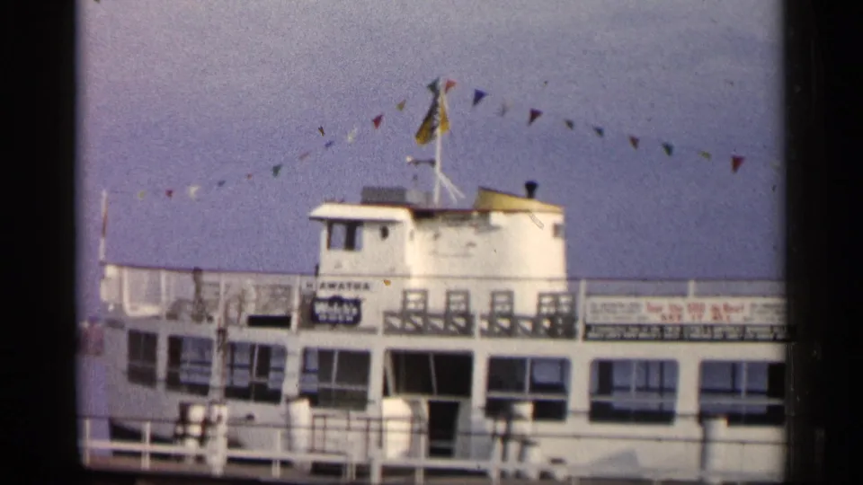 1960 Michigan home-movie frame of a white docked passenger boat with railings, signage, and festive bunting overhead.