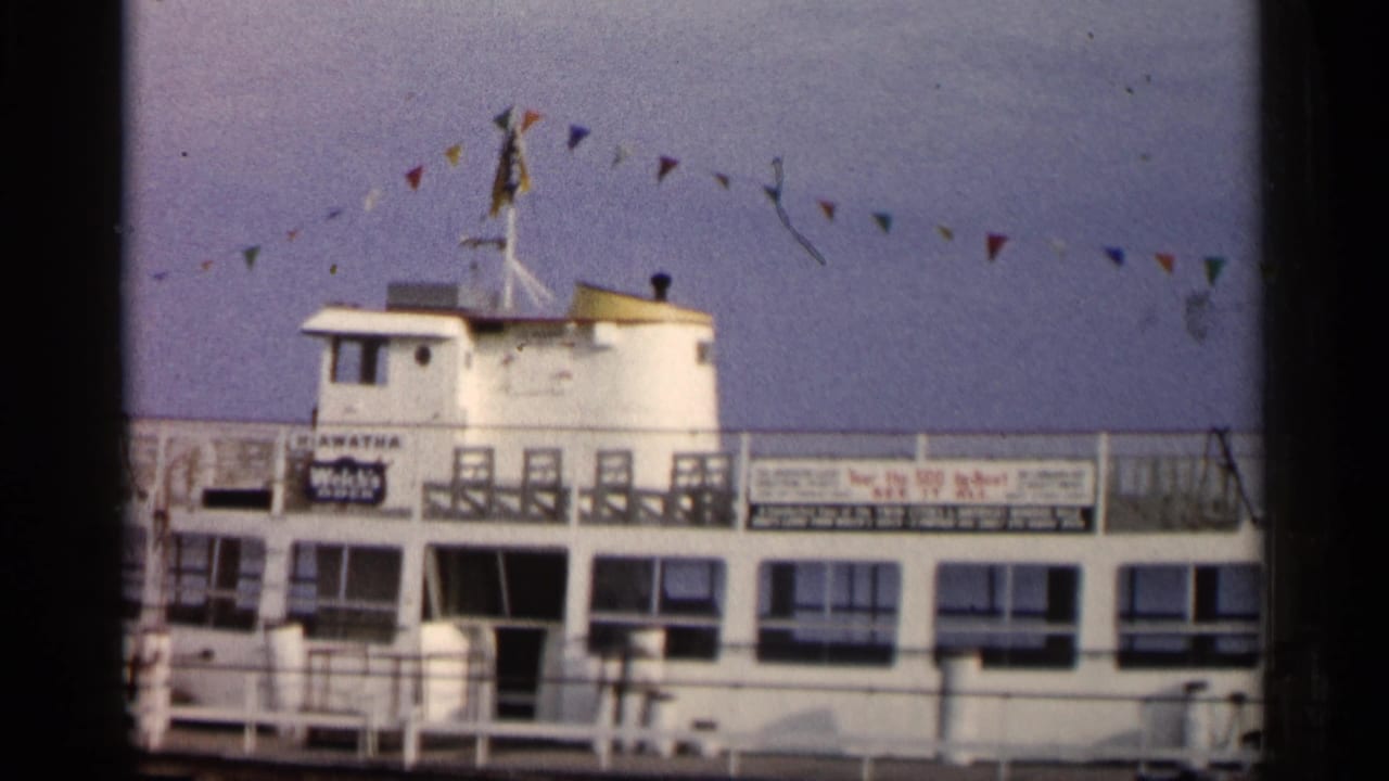 Broadside 1960 Michigan waterfront view of a white passenger boat, upper railings, readable vessel signage, and colored bunting.