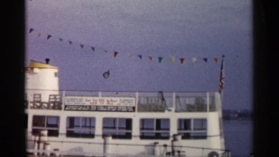 1960 Michigan home-movie frame of the right side of a docked passenger boat with bunting, signage, calm water, and an American flag.