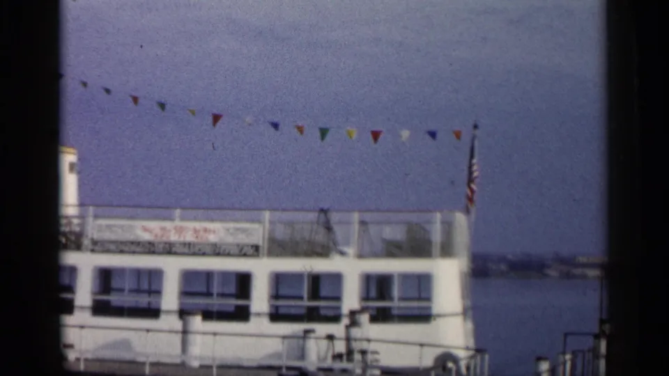 1960 Michigan 8mm frame showing a white tour boat at a dock with pennant flags and a river behind it.