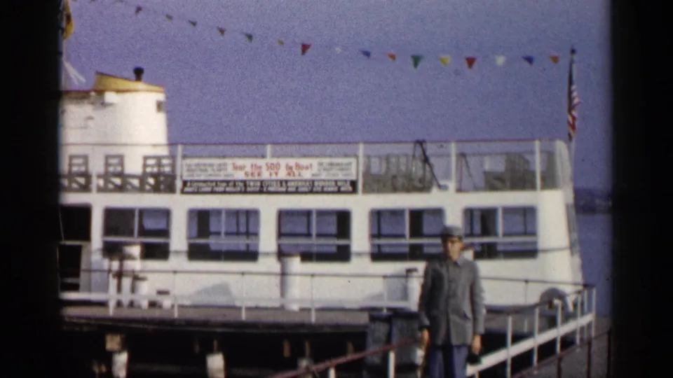 1960 Michigan 8mm frame with a man on the dock in front of a white tour boat, railings, flags, and signage.