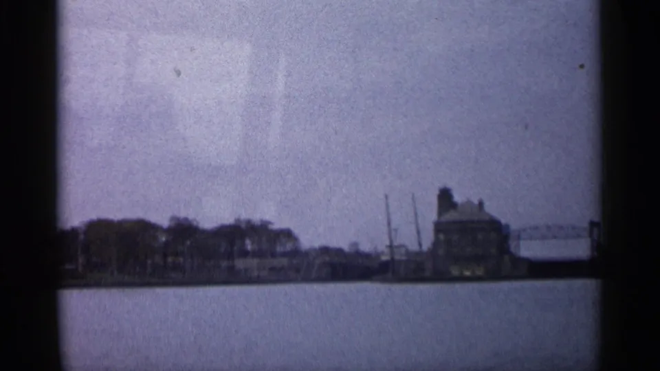 1960 Michigan 8mm frame showing a distant shoreline, low structures, and calm river water from a boat or dockside viewpoint.