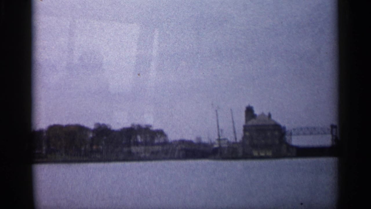 1960 Michigan home-movie frame showing a distant shoreline, calm water, trees, and a large building near a dock area.
