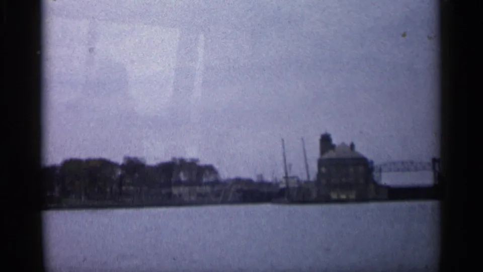 A soft 8mm view across water toward a Michigan waterfront building, trees, and dockside structures in 1960.
