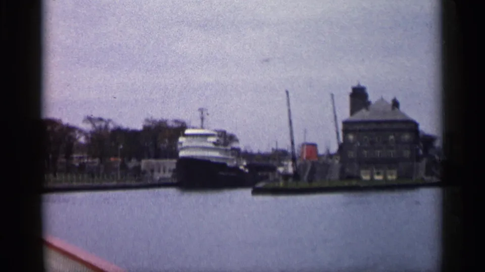 1960 Michigan waterfront frame with a large vessel near a pier, a dockside building, trees, and calm water in the foreground.
