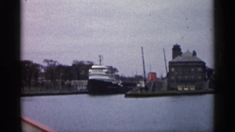 The vessel sits near a pier on a quiet 1960 Michigan waterfront, with a large building and vertical dock structures behind it.
