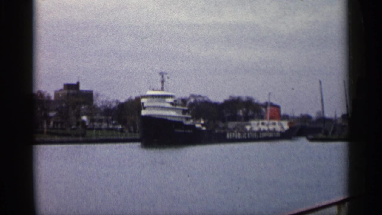 1960 Michigan harbor view of a large freighter moving past a small craft and low industrial shoreline.