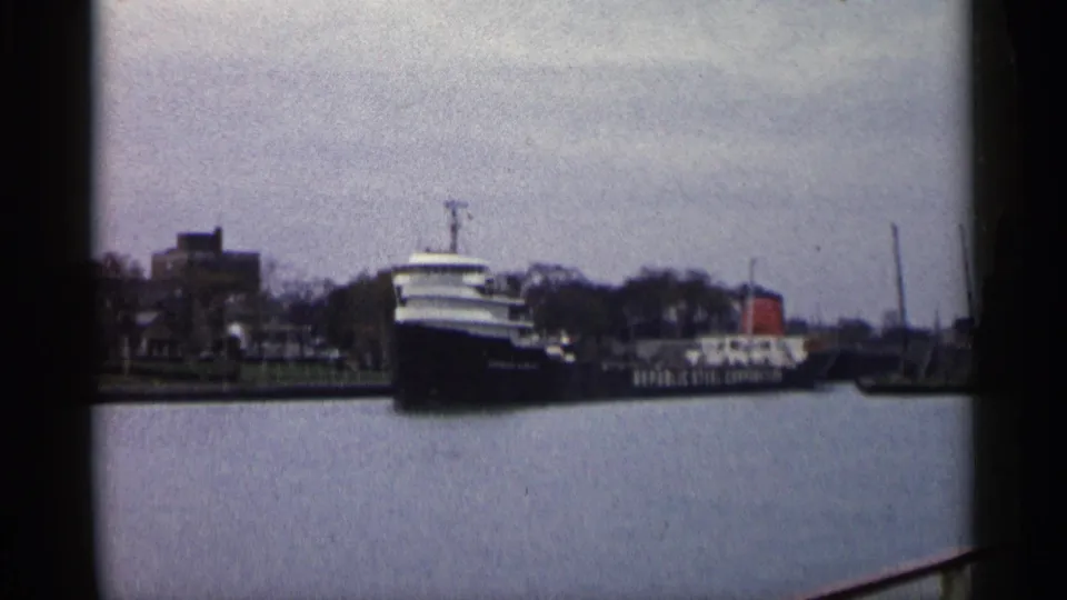 1960 Michigan harbor view of a large freighter moving past a small craft and low industrial shoreline.