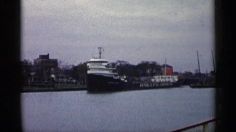 1960 Michigan 8mm frame of a dark-hulled freighter with visible side lettering passing a working waterfront.