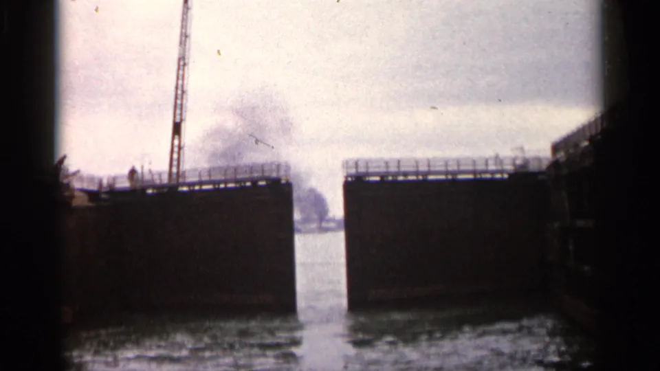 1960 home-movie frame from a boat inside or near the lock, showing dark walls, a crane, open water, and soft film grain.