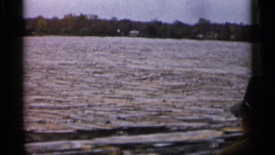 Michigan 8mm frame looking across choppy water toward a wooded shoreline, with a boat rail and partial hatted passenger at the right edge.
