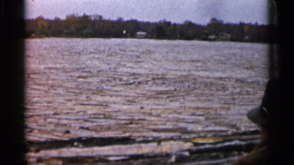 1960 Michigan 8mm frame from a boat, showing rippled river water, distant shore buildings, trees, and a hat-wearing passenger silhouette.
