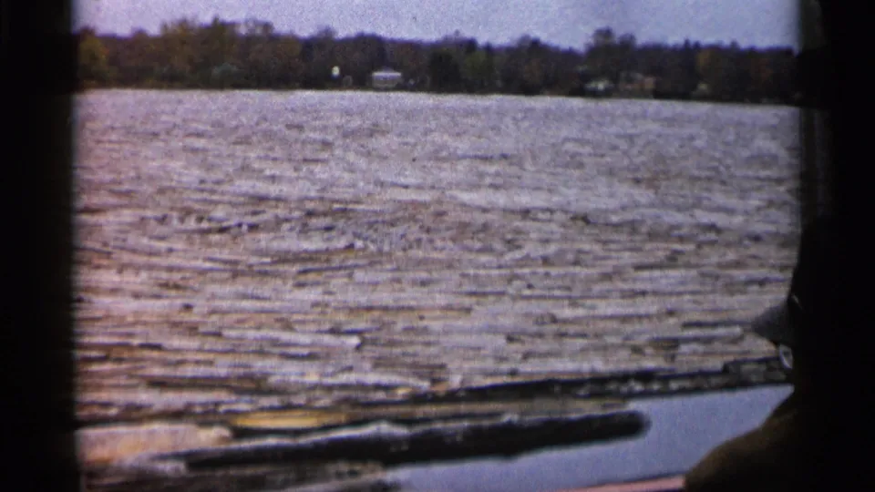 1960 Michigan boat-view frame with rippled water in the foreground, a low shoreline, distant buildings, and a partial passenger silhouette.