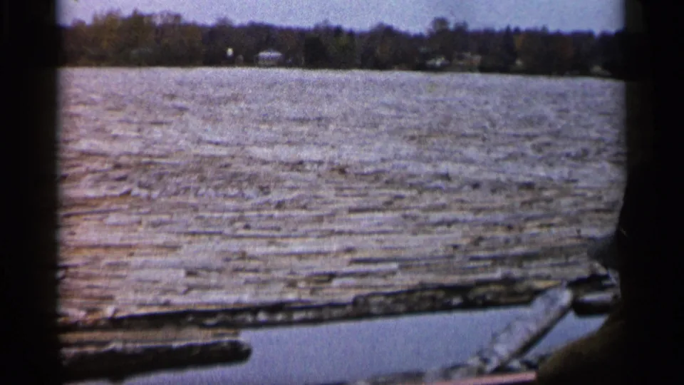 1960 Michigan 8mm river view from a moving boat, with textured water, distant trees, shoreline buildings, and a passenger silhouette.