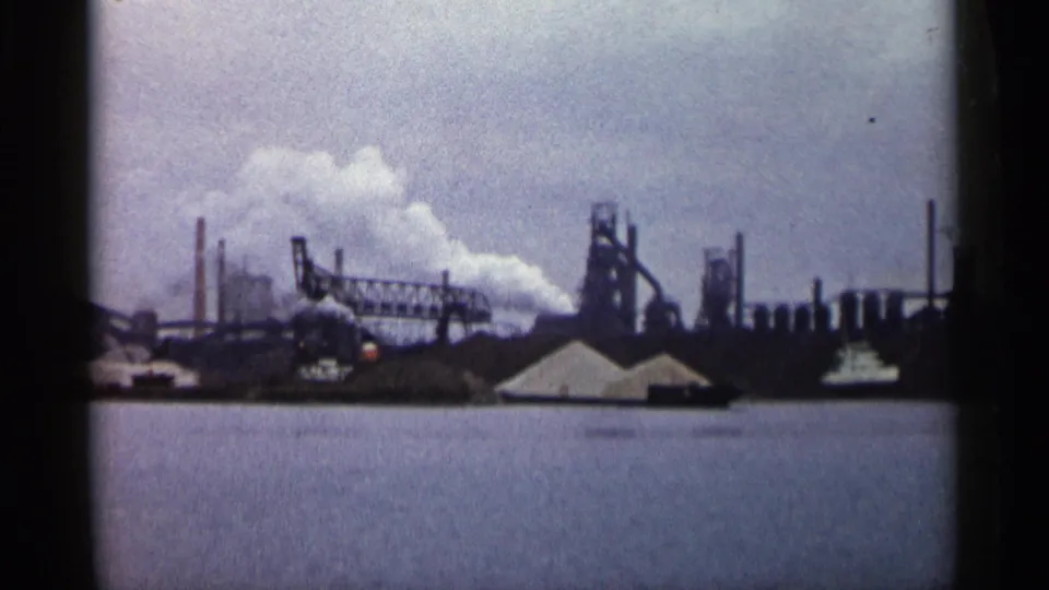 1960 Michigan 8mm frame showing an industrial complex with smokestacks, smoke plumes, low buildings, and water in the foreground.