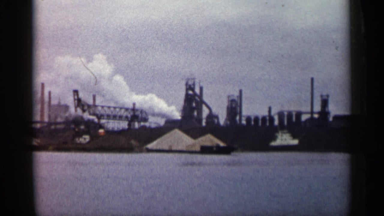 Factory skyline with smoke plume and water in the foreground.